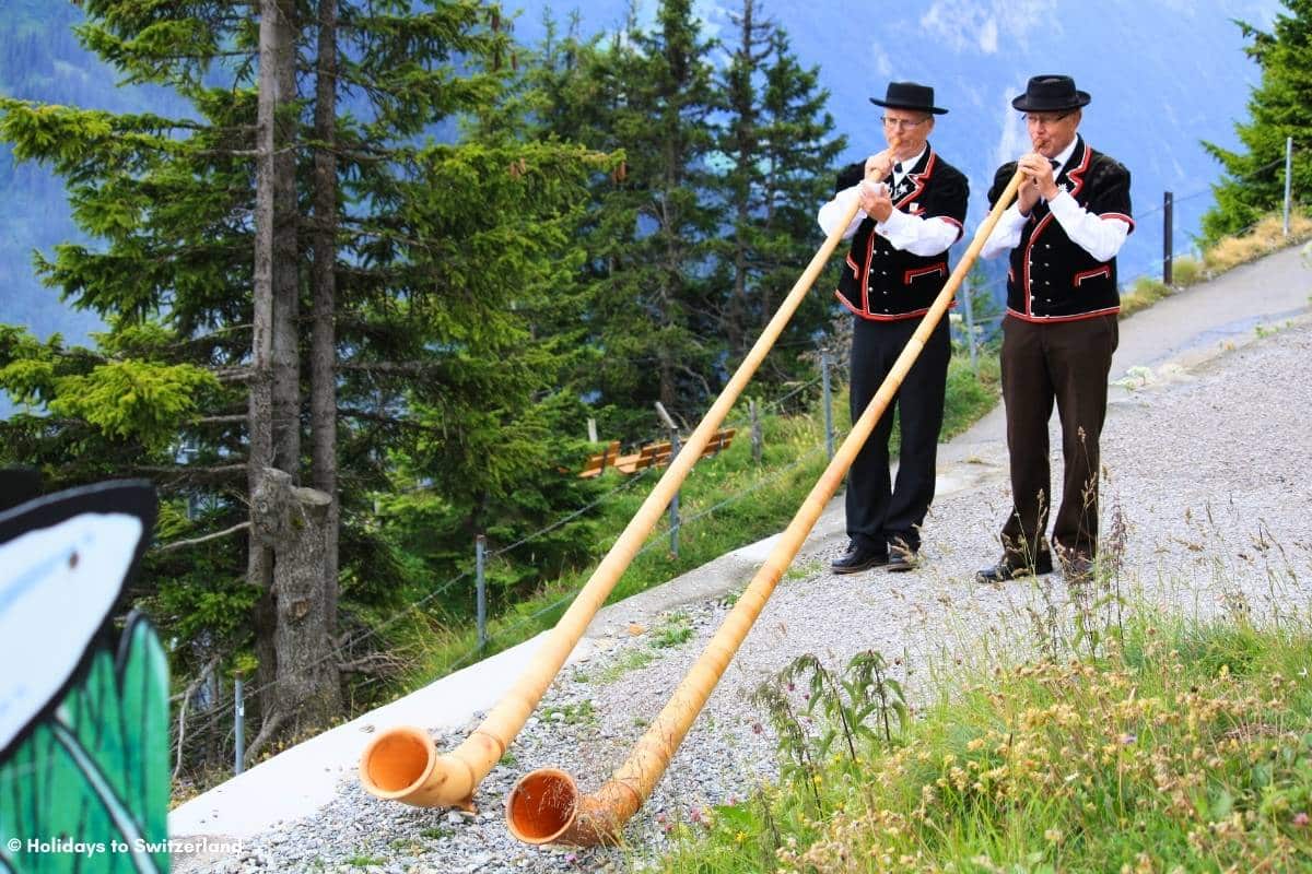 Two men dressed in traditional Swiss costumes whilst playing Alphorns