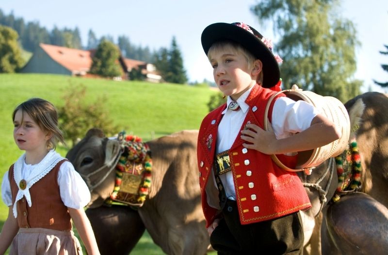 Two children dressed in traditional Appenzell costumes walking beside some cows.