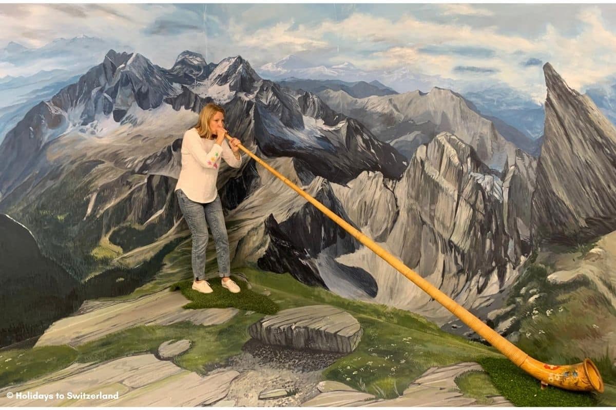 Woman playing an Alphorn in front of a painted mural of a mountain scene