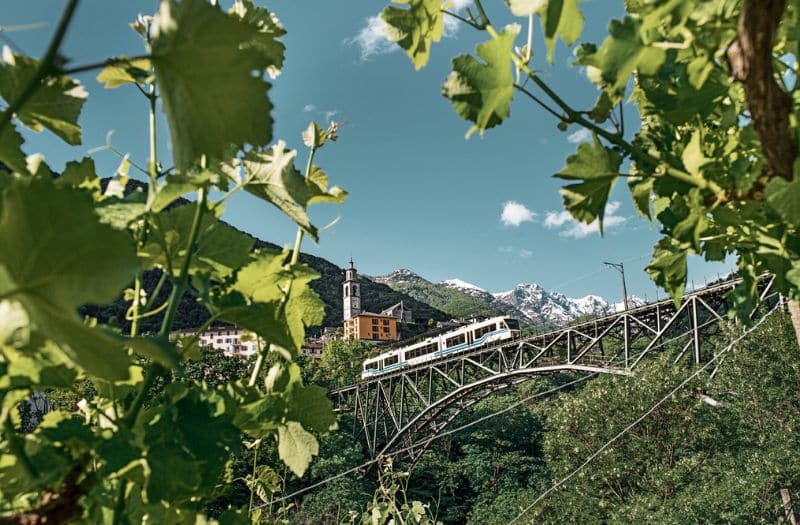 A train of the Centovalli Railway crossing a steel bridge with a mountain backdrop