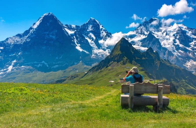 Man seated on a bench whilst taking a photo of the Eiger, Monch and Jungfrau from Mannlichen