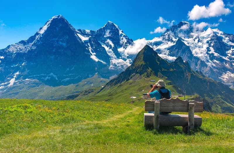 Man seated on a bench whilst taking a photo of the Eiger, Monch and Jungfrau from Mannlichen