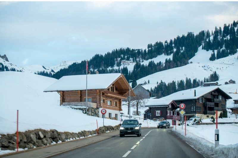 Cars drive on a cleared road through a village which is covered with snow