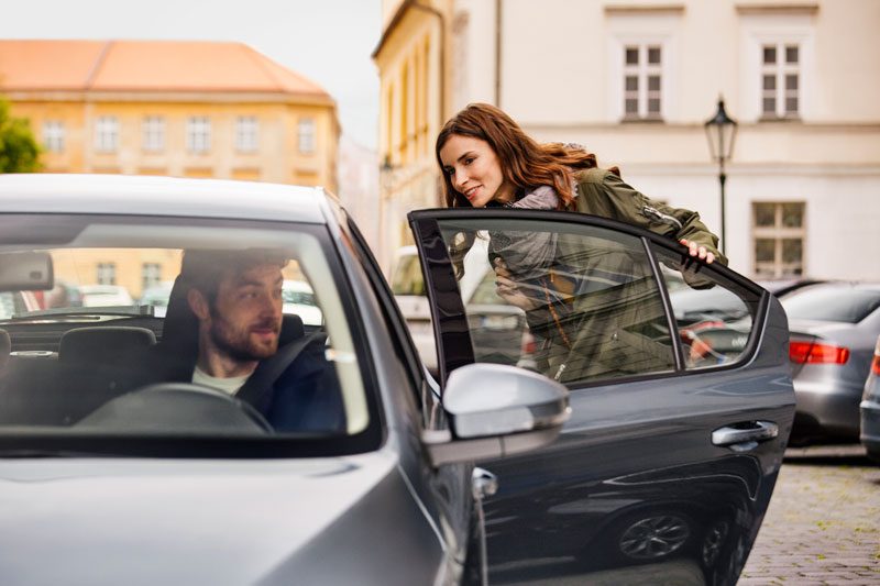 A woman about to sit in the back of a vehicle whilst the driver looks on from the front seat.